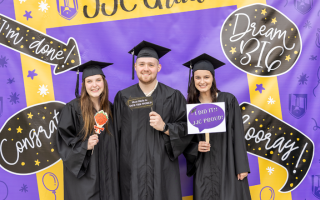 three people pose for photo with props in graduation attire in front of graduation backdrop