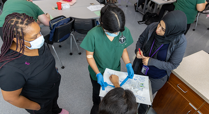 three medical assisting students practice in the classroom with their professor observing