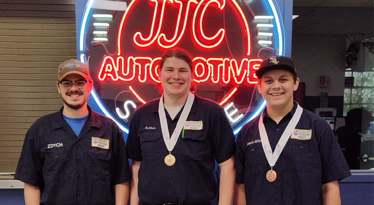three people (two wearing medals) pose for photo in front of JJC Automotive neon light