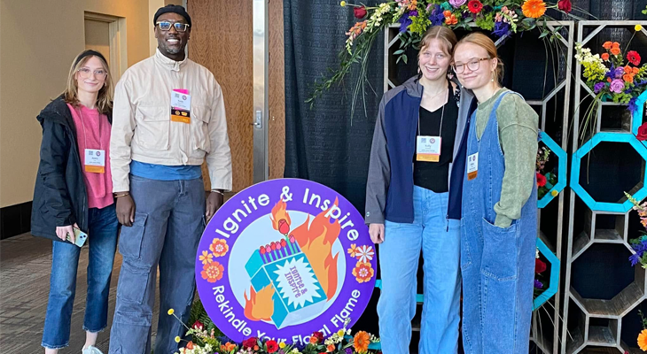 four people pose for photo next to floral arrangement on ground with circular sign that says