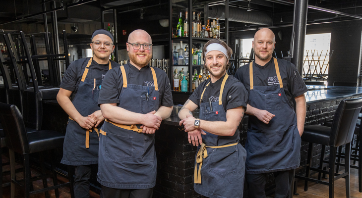 four chefs pose for photo in restaurant while wearing matching black outfits and aprons