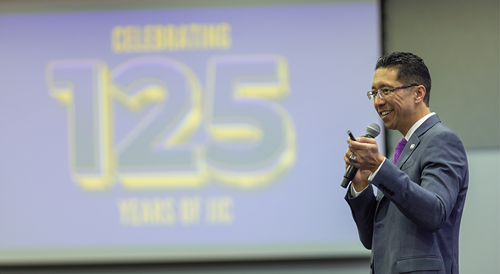 Clyne Namuo stands and speaks into microphone with projection display behind him blurred in background that says
