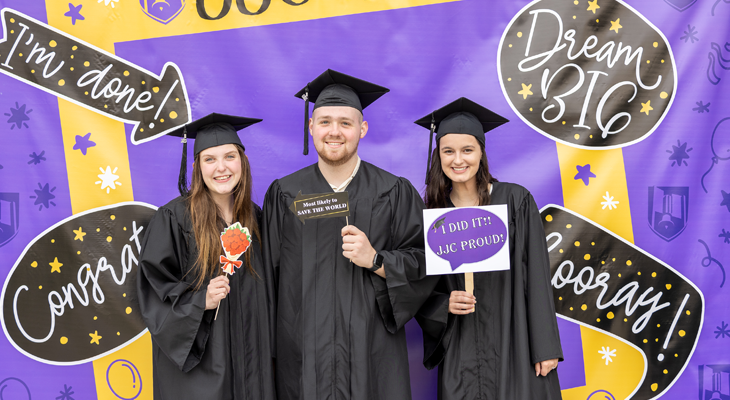 three graduates pose for photo and hold props in front of graduation backdrop