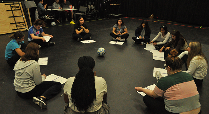 cast members of play sit in circle on floor during rehearsal